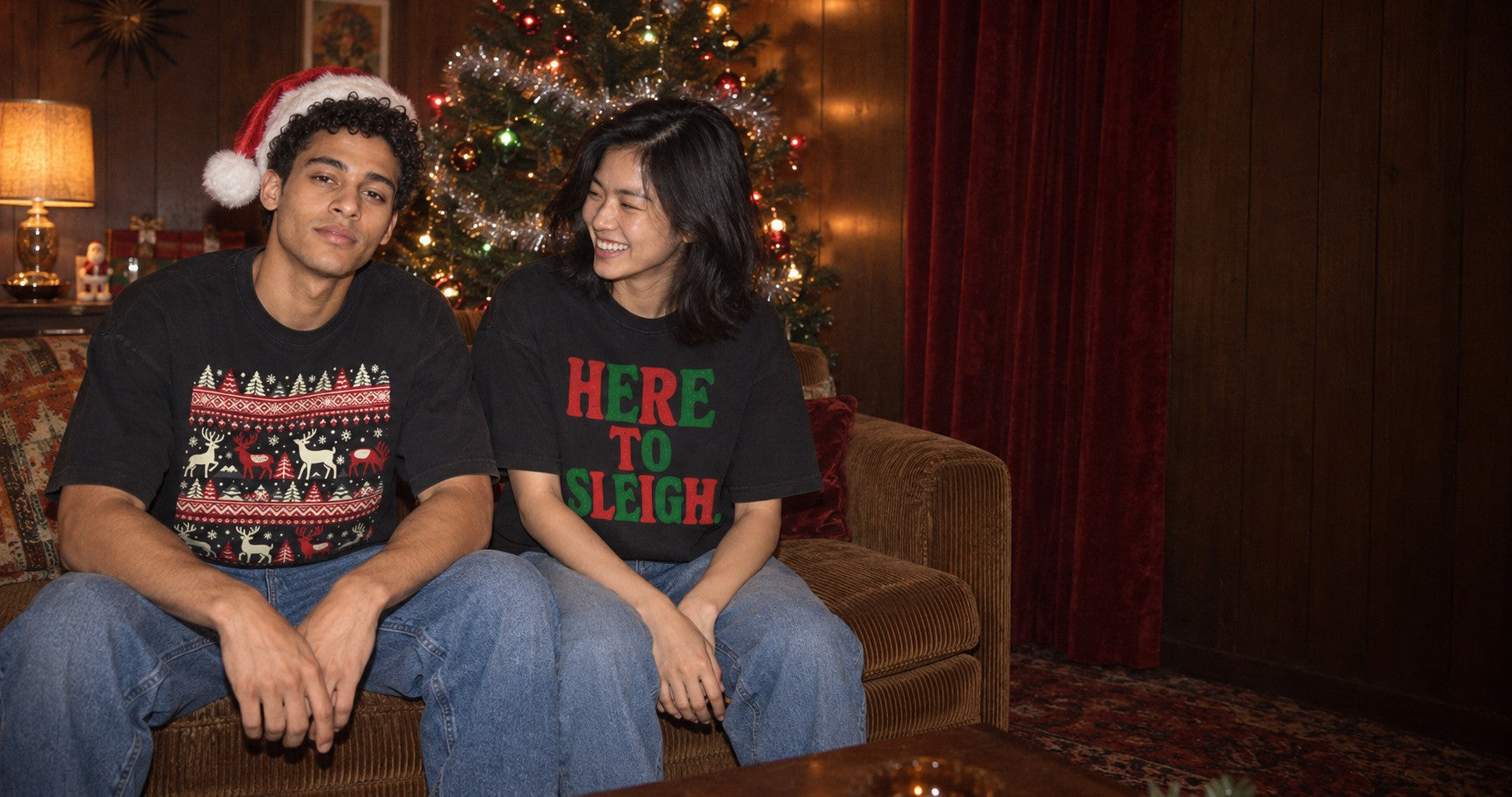 Two friends on a sofa beside a Christmas tree in Teekop tees printed with a reindeer pattern and the words Here to Sleigh