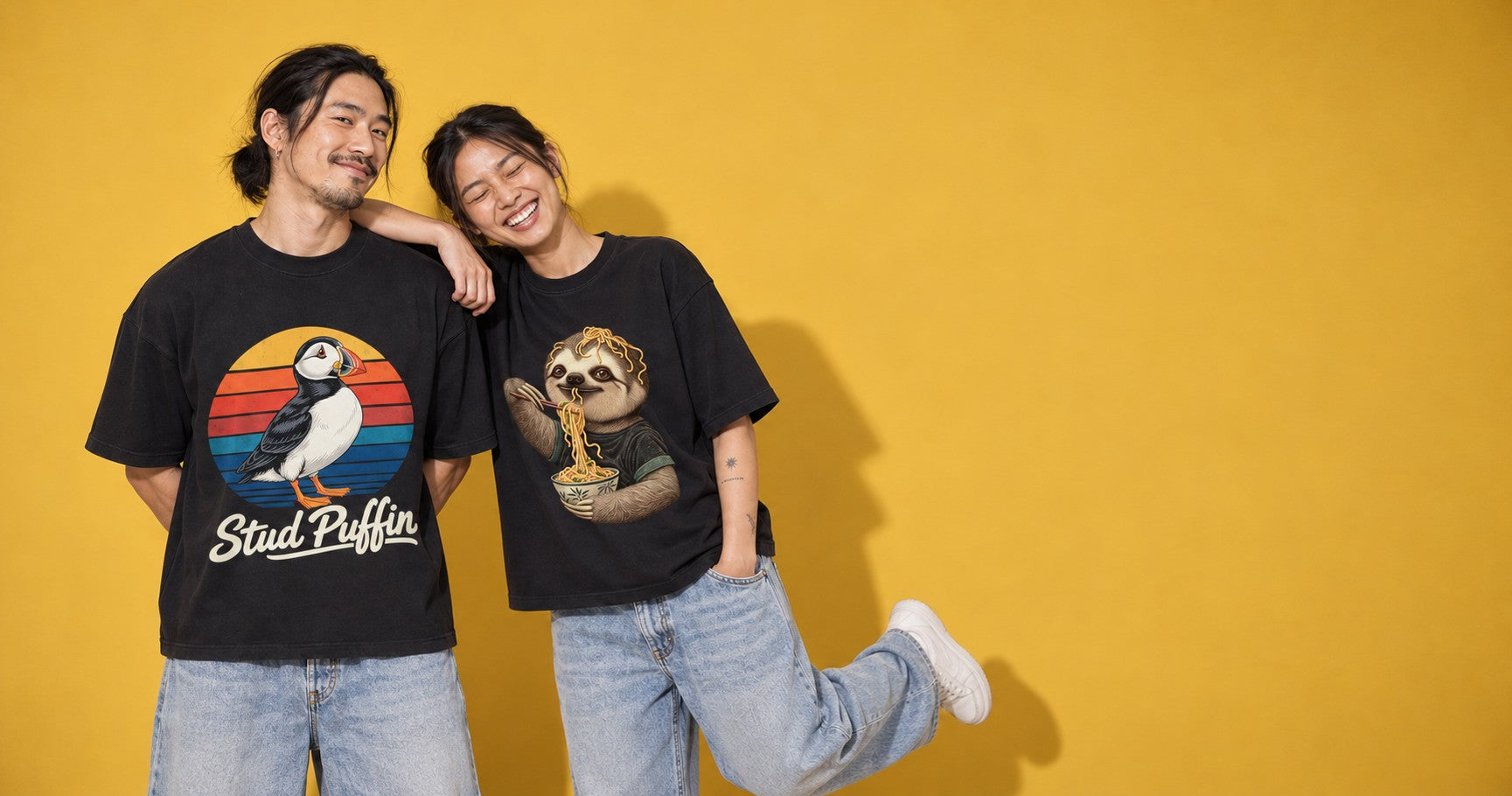 Two friends laughing in front of a yellow backdrop in Teekop tees printed with a puffin and a sloth eating noodles